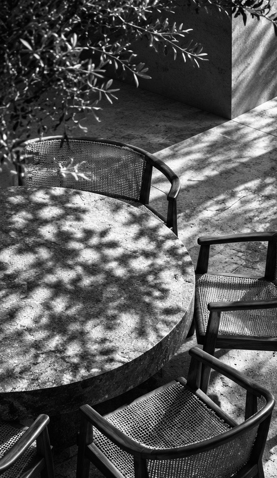 A round stone table and woven chairs under dappled shade.